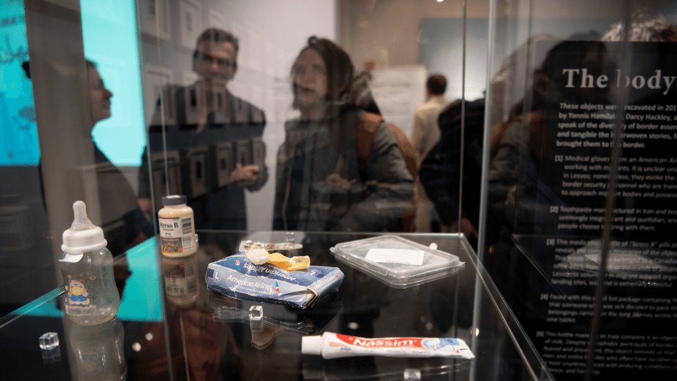 A baby bottle, toothpaste, and box of gloves on display in a glass case, with visitors visible in the case reflection.