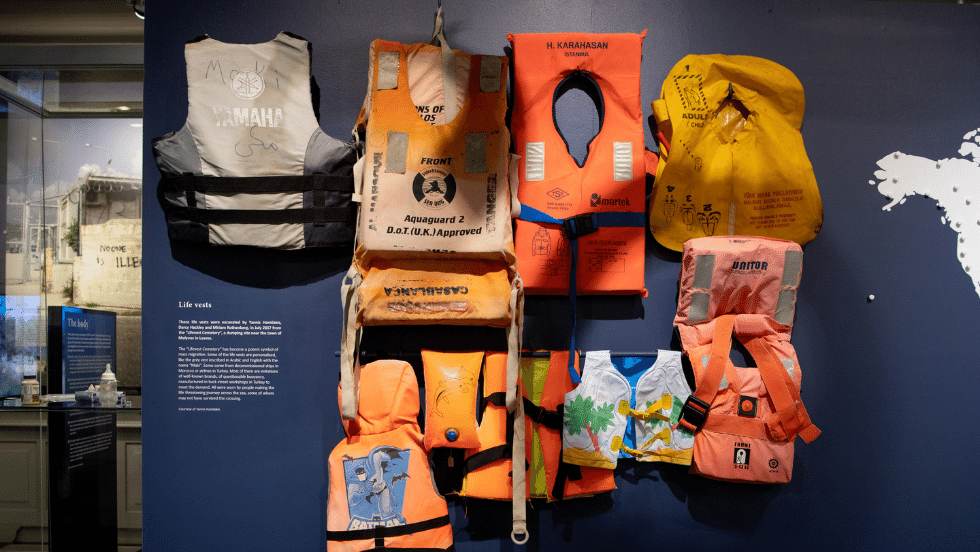 Different sized and colored life-vests hung on the wall beside an exhibit label. 