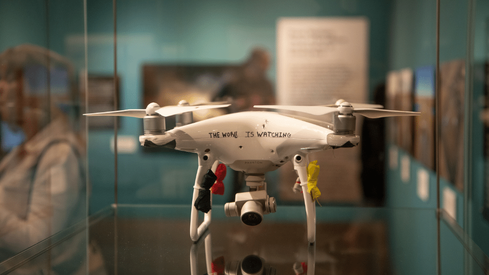 Photo of a drone in an exhibit case, with "the world is watching" written on it.