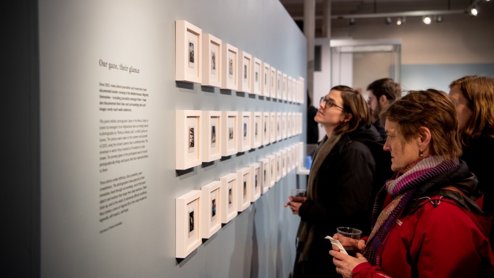 Photo of visitors at the opening of the Transient Matter exhibit looking at a wall of framed polaroids. 