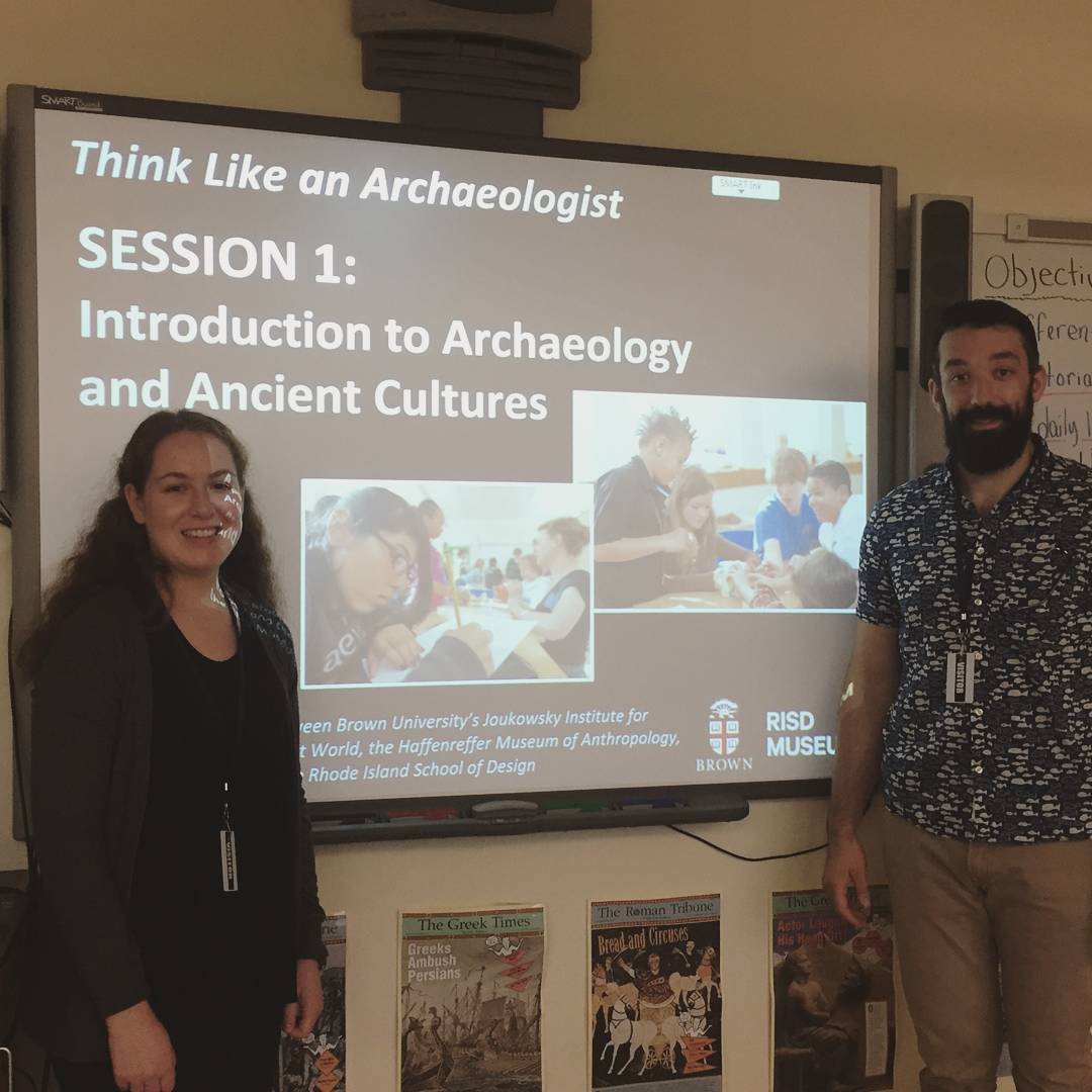 Two Brown University students standing in front of projector screen with the Think Like An Archaeologist presentation, in an elementary school classroom.