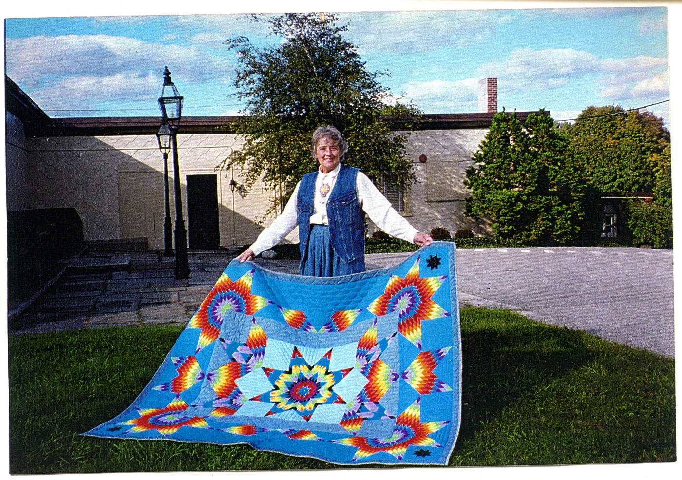 Photo of Barbara Hail, white woman with short hair, holding a large patchwork blanket in front of the Haffenreffer Museum in Bristol, RI.