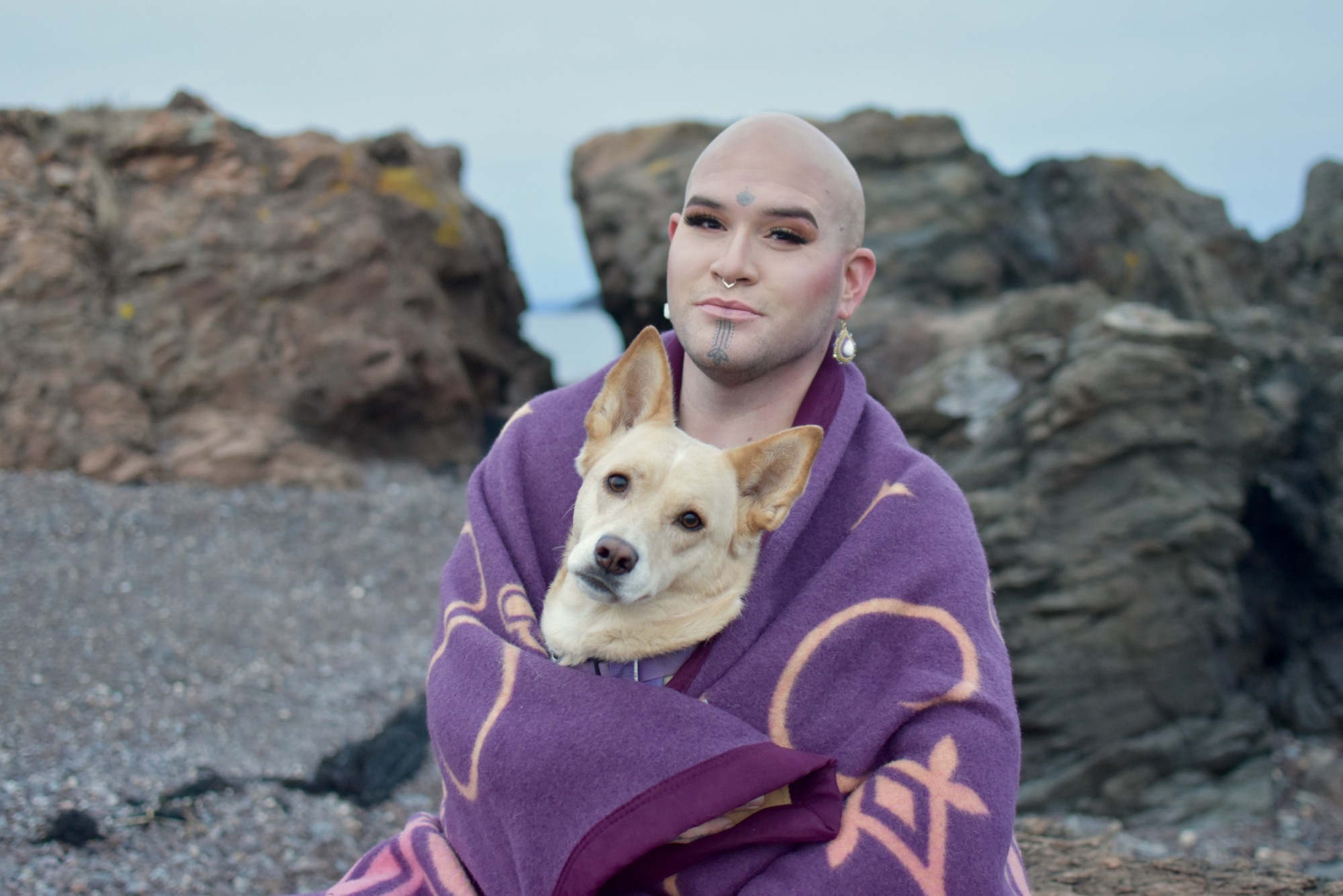 Photo of Geo Neptune sitting on rocks at the beach with their dog and wrapped in a purple blanket. 