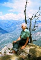 Photo of Gino E. Conti sitting on a rock overlooking a mountainous landscape. 