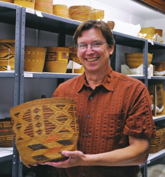 Photo of Robert Preucel holding a woven basket, standing in collection storage.