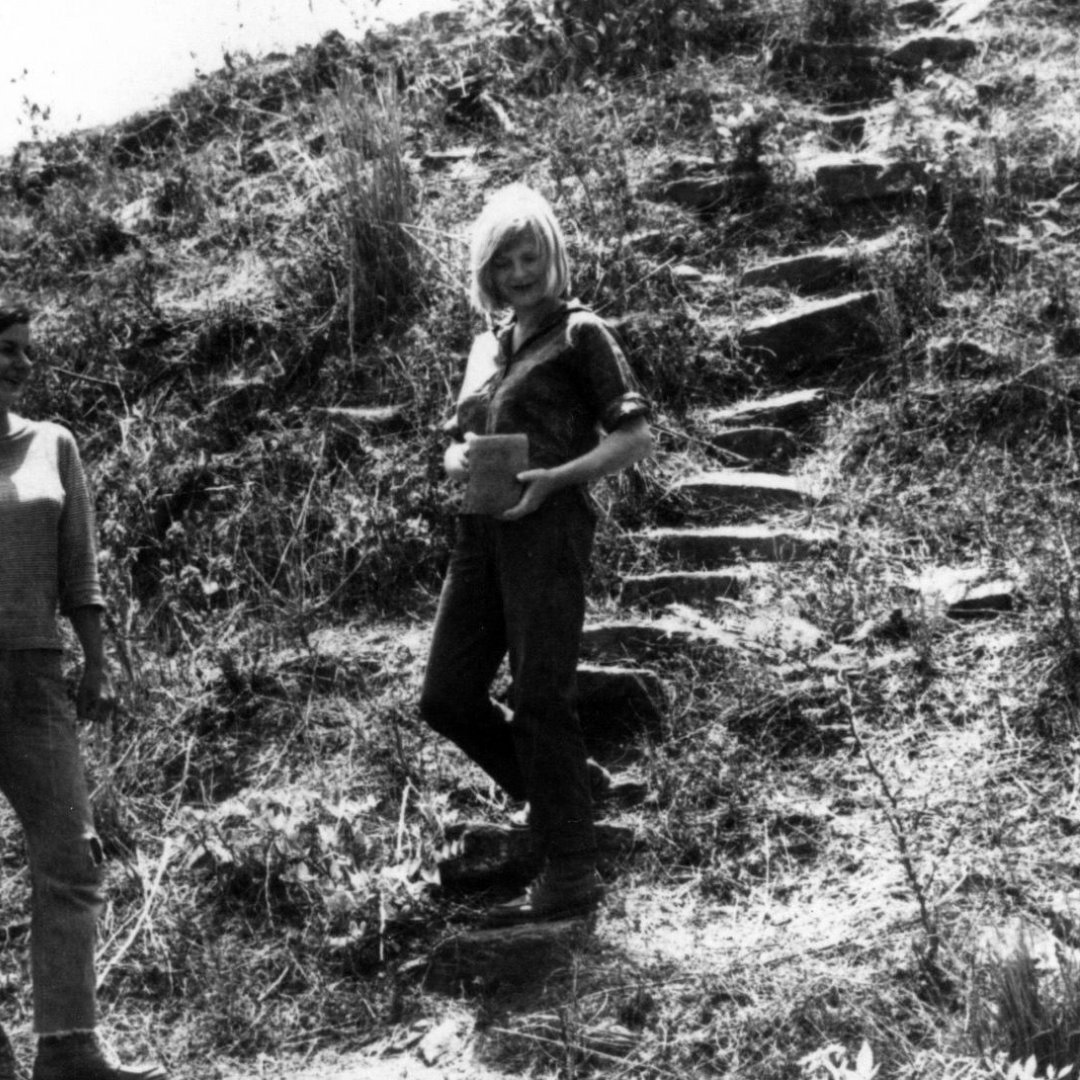 Black and white vintage photo of Jane Dwyer walking down an embankment holding a pot.