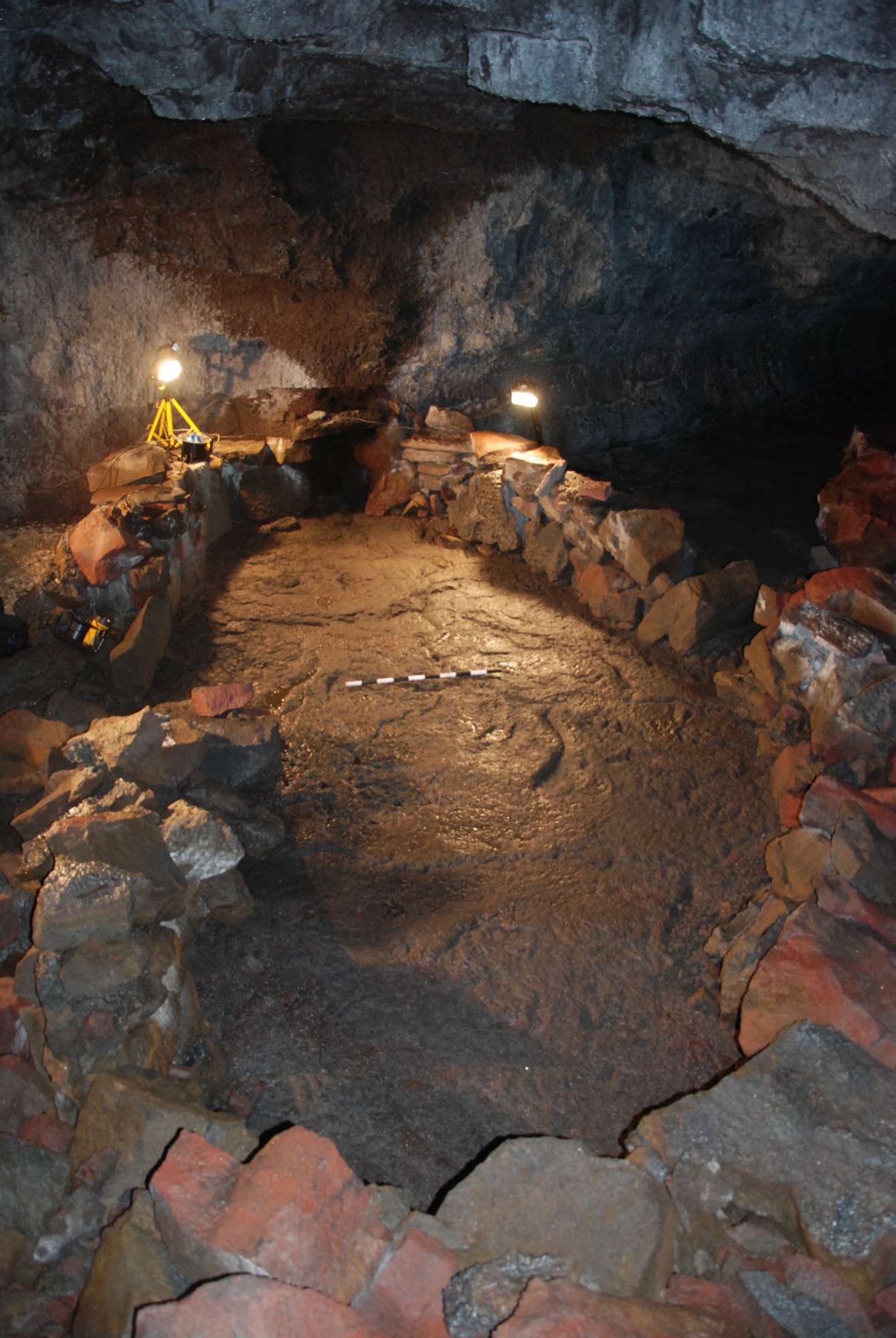 Photo of the dark interior of a large cave. A ring of stones surrounds a smooth stone floor illuminated with two lamps. 