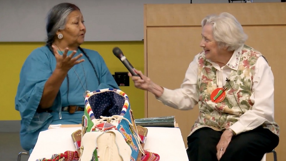 Two woman seated in front of a beaded cradleboard, one Native American woman in a blue dress and one white woman in a white and floral shirt holding out a microphone. 